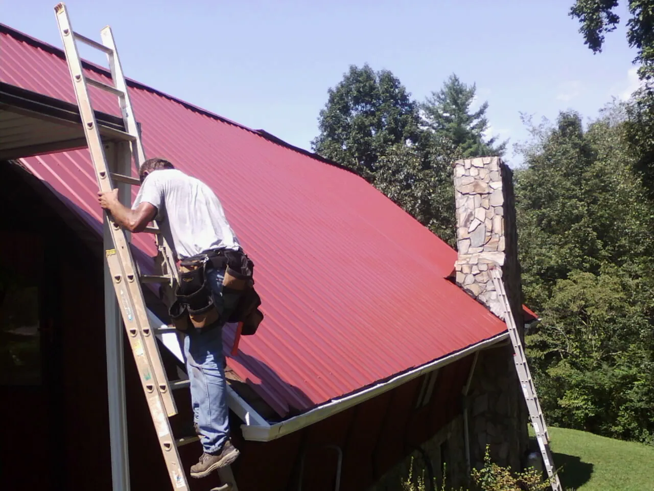 Roofer on a ladder beside a red metal roof and stone chimney — Soffit & Fascia Repair by Roofing Brothers in Antigo